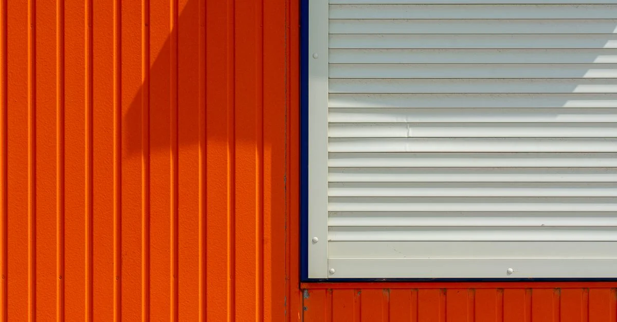 Mur orange minimaliste avec une fenêtre à volets blancs, mettant en valeur de fortes lignes géométriques et textures.