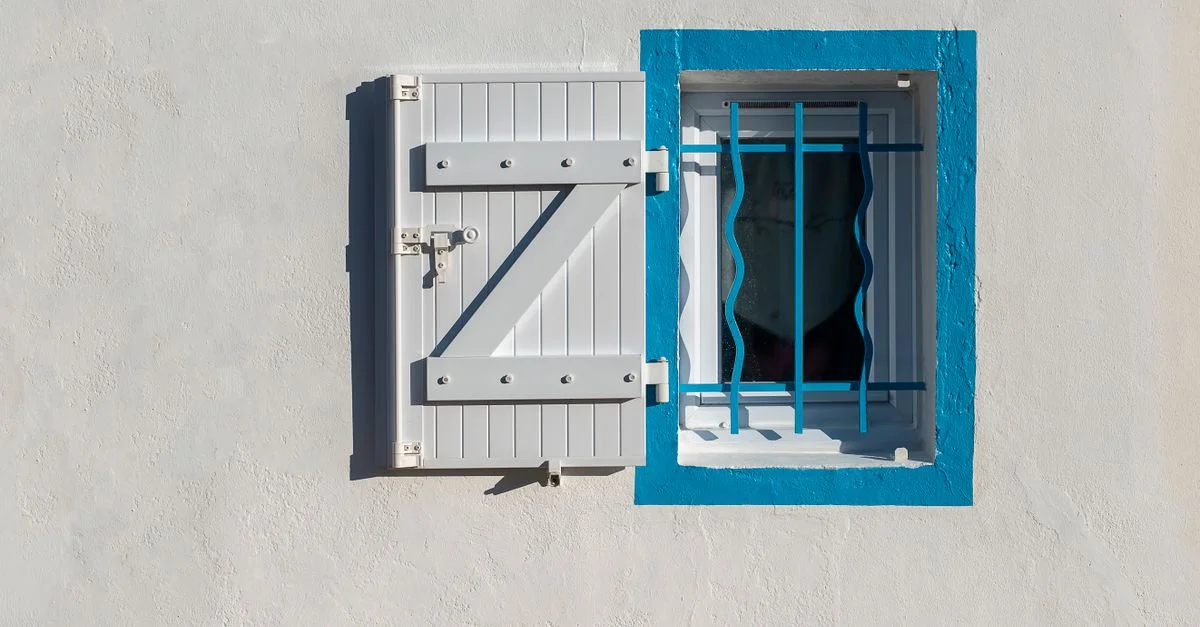 Fenêtre blanche et bleue avec volets sur un mur ensoleillé, mettant en valeur un design architectural minimaliste.