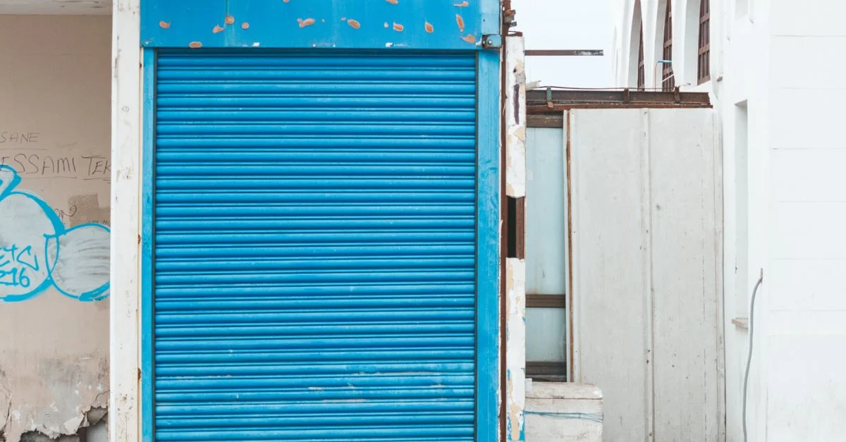 Un volet roulant bleu sur une façade de bâtiment en décomposition, affichant des graffitis et des signes de dégradation.
